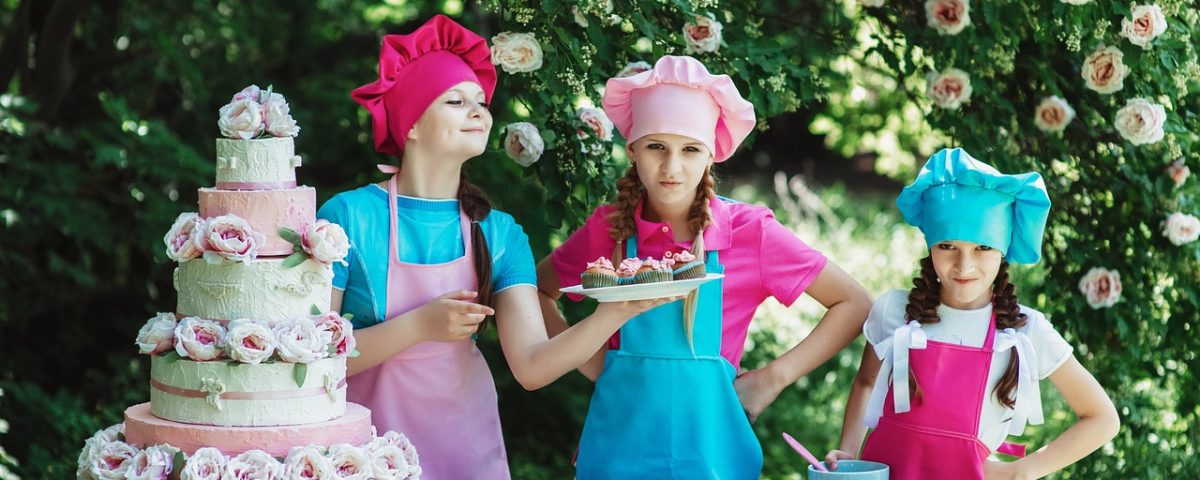 kids in baking aprons and hats alongside table with cake